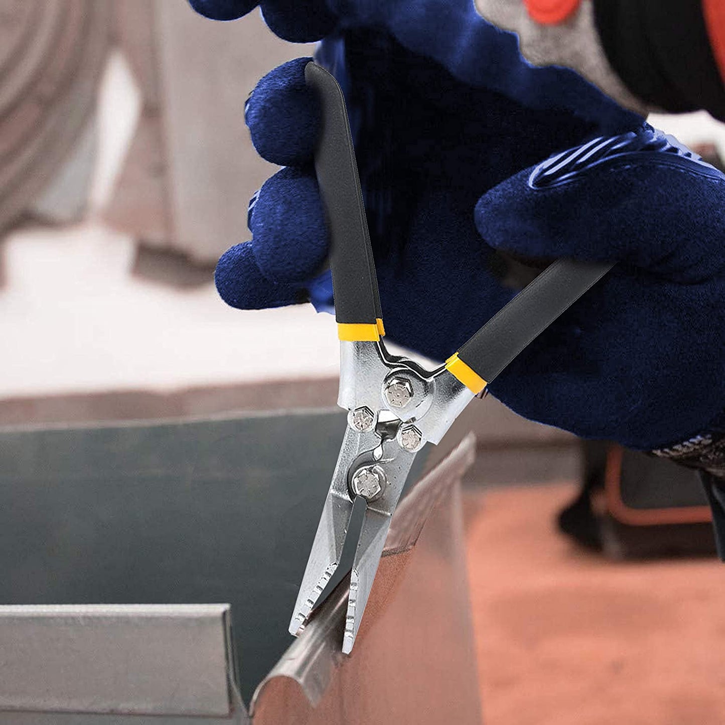A person wearing blue gloves is using a pair of High Quality Sheet Metal Hand Seamers - Multiple Sizes Available by Perma Cover to bend a metal sheet. The heavy-duty all-steel pliers have black handles with yellow accents. The background includes blurred industrial equipment and fabrics.