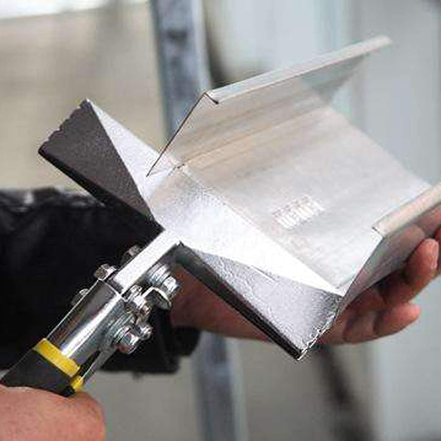 A close-up of a person using Perma Cover High Quality Sheet Metal Hand Seamers - Multiple Sizes Available to cut or shape a piece of metal. The tool has a yellow and black handle, perfect for bending, seaming, and flattening the shiny, angular metal piece. The background is blurred with indistinct shapes and colors.
